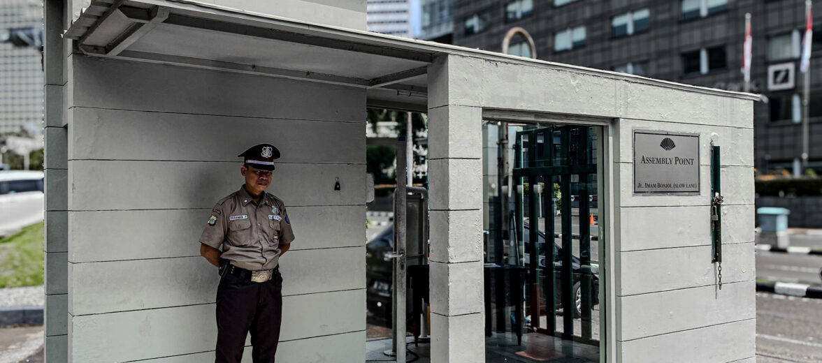 Petugas keamanan berpakaian seragam berdiri di pos jaga di depan sebuah gedung perkantoran, melambangkan keamanan fisik, dengan latar belakang urban yang mencerminkan kebutuhan untuk keamanan digital yang terintegrasi dalam lingkungan bisnis modern.