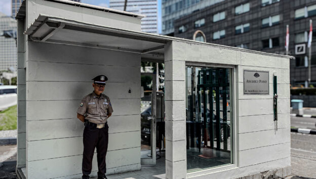 Petugas keamanan berpakaian seragam berdiri di pos jaga di depan sebuah gedung perkantoran, melambangkan keamanan fisik, dengan latar belakang urban yang mencerminkan kebutuhan untuk keamanan digital yang terintegrasi dalam lingkungan bisnis modern.