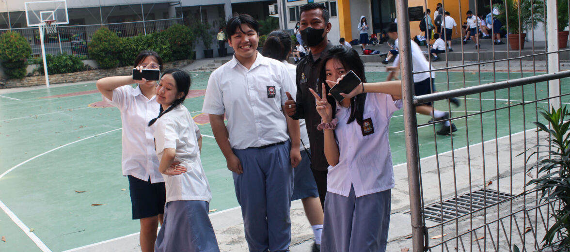 Seorang petugas keamanan berdiri bersama siswa yang sedang bersantai di lingkungan sekolah, menunjukkan keamanan lingkungan pendidikan yang terjaga.