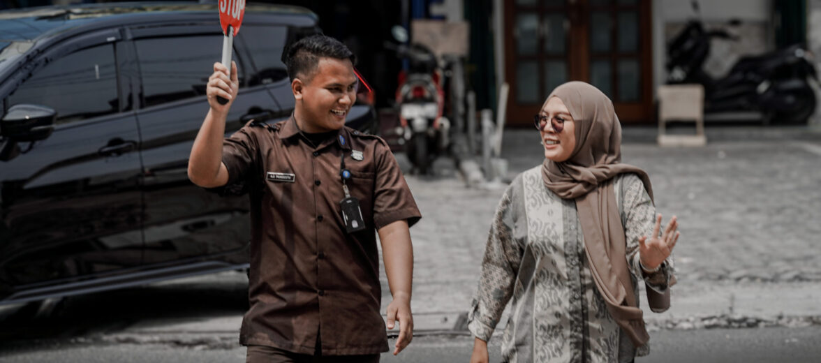 Seorang petugas keamanan berseragam cokelat dengan ID terlihat sedang mengarahkan lalu lintas dengan menggunakan papan STOP sambil tersenyum kepada seorang wanita berhijab yang juga tersenyum. Keduanya berjalan bersama di area parkir yang tampak tenang dan teratur, menandakan lingkungan kerja yang aman dan terkendali.