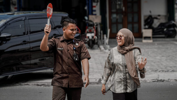 Seorang petugas keamanan berseragam cokelat dengan ID terlihat sedang mengarahkan lalu lintas dengan menggunakan papan STOP sambil tersenyum kepada seorang wanita berhijab yang juga tersenyum. Keduanya berjalan bersama di area parkir yang tampak tenang dan teratur, menandakan lingkungan kerja yang aman dan terkendali.